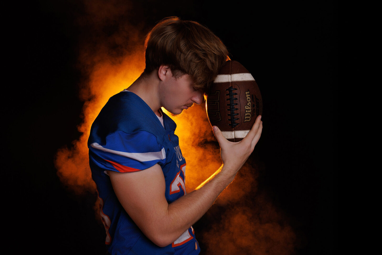 High school football player holding a football with vibrant orange smoke behind him during a dramatic sports photography session.