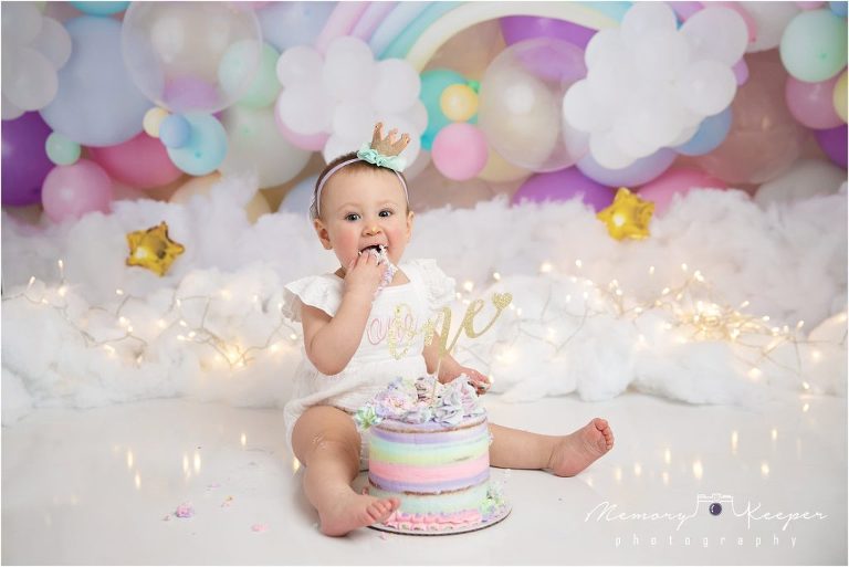Baby girl eating a rainbow colored cake with a rainbow/dreamy theme backdrop. 